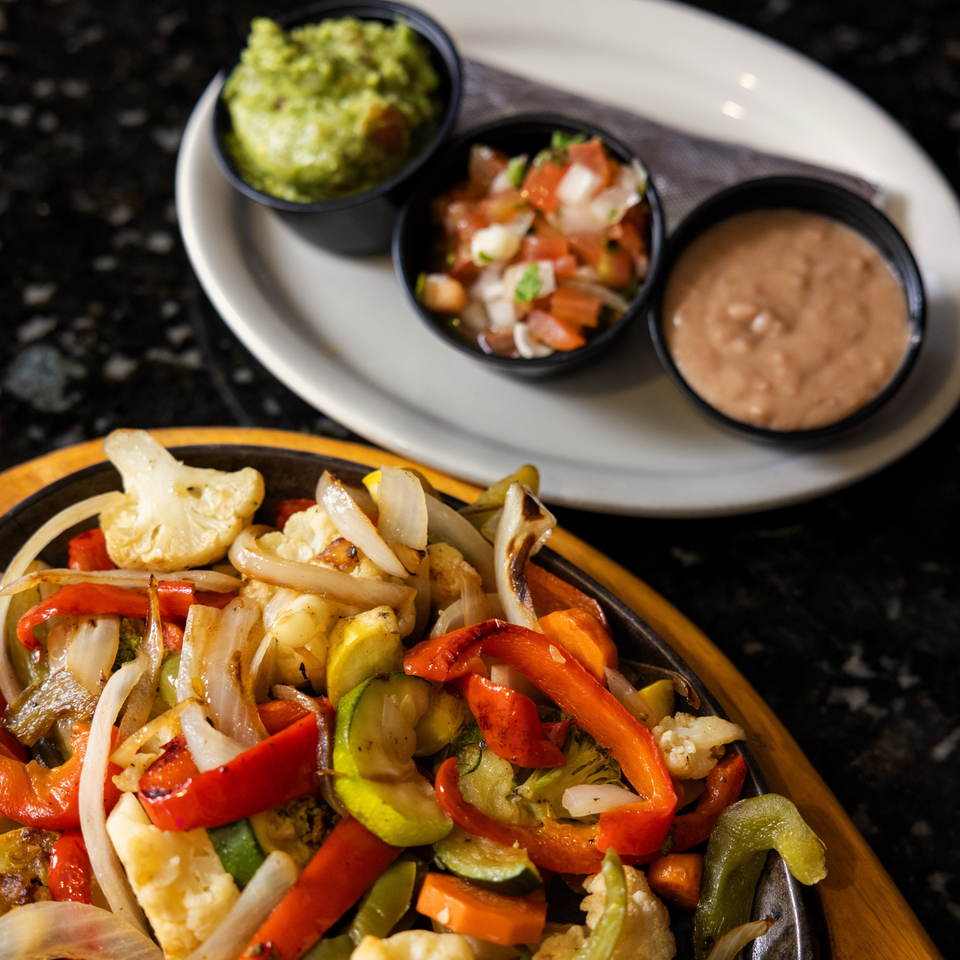 Sizzling fajita vegetables with sides of guacamole, pico de gallo, and refried beans.