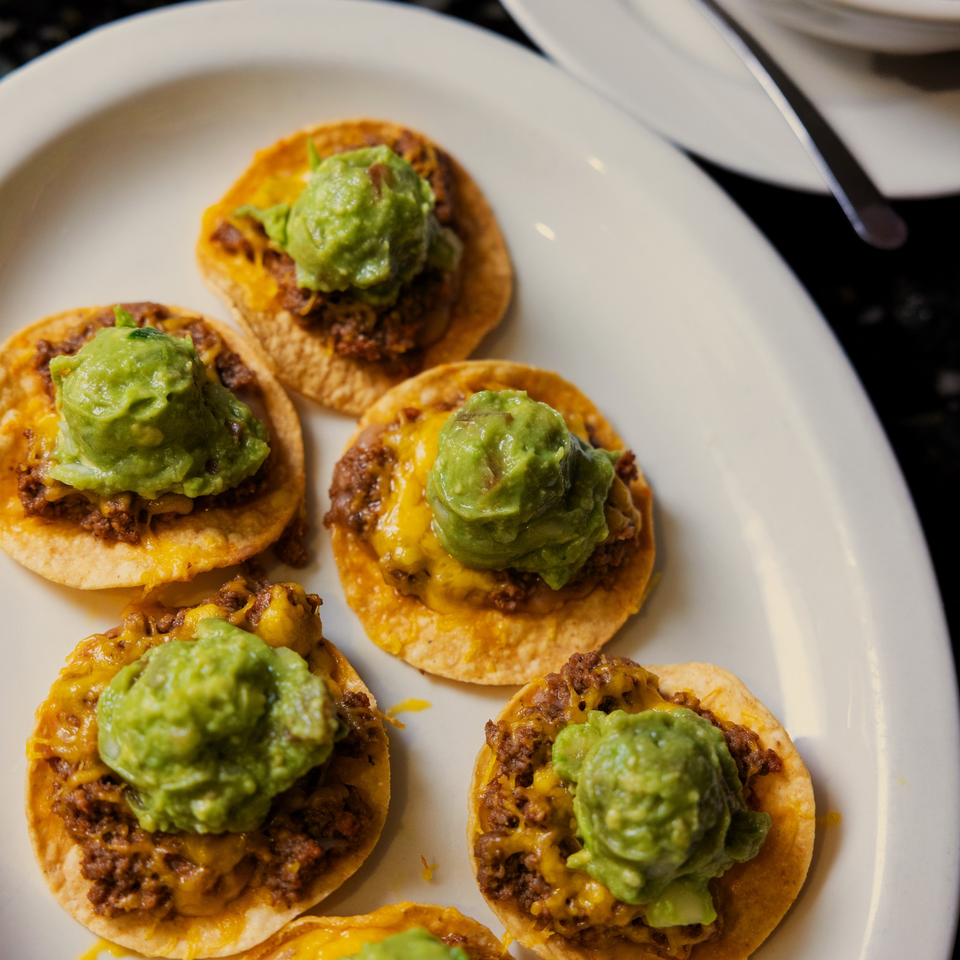 A white plate holds several mini tostadas topped with ground beef, melted cheese, and guacamole.