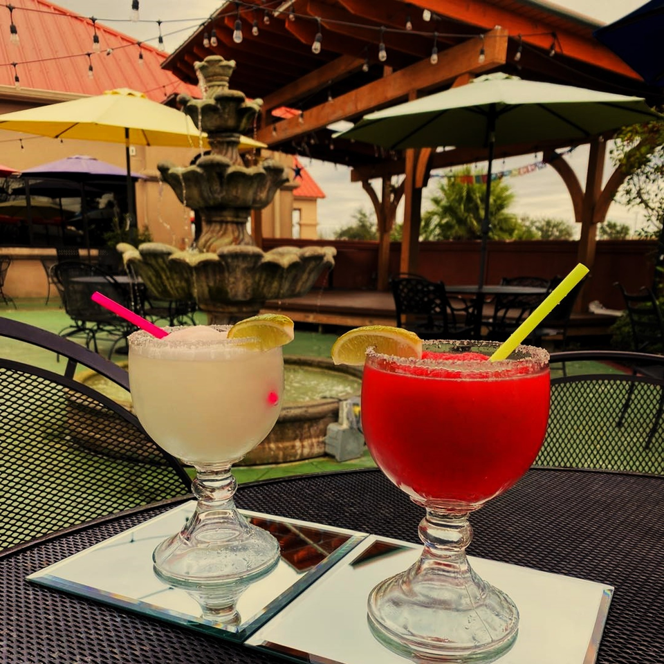 Two margaritas, one red and one white, on a patio table with a fountain and umbrellas.