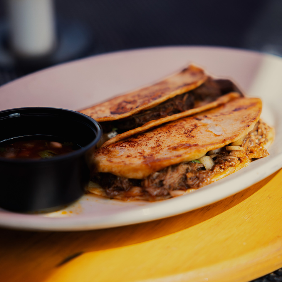 Two La Casa del Sol birria tacos with a side of consome on a white plate.
