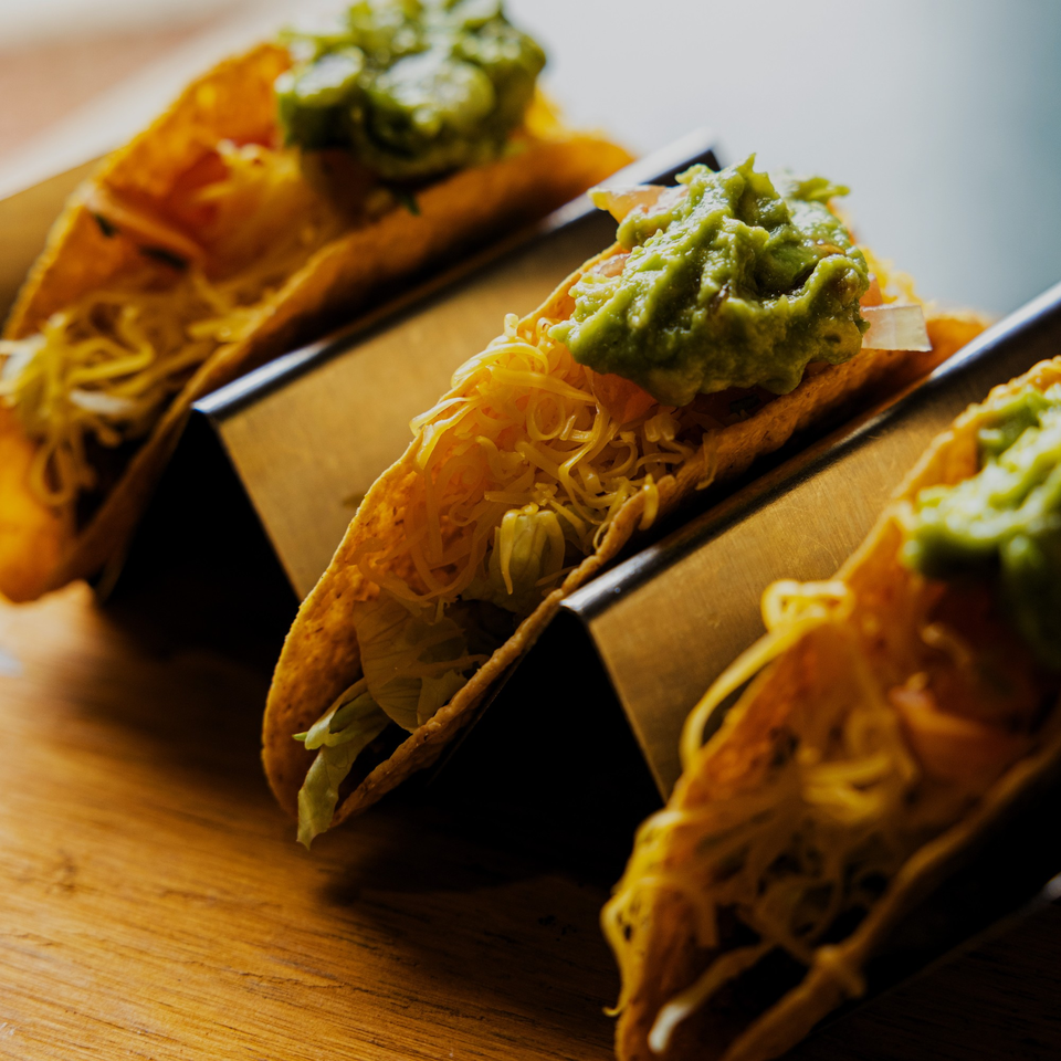 Three La Casa del Sol tacos with guacamole, shredded cheese, and lettuce in metal holders.